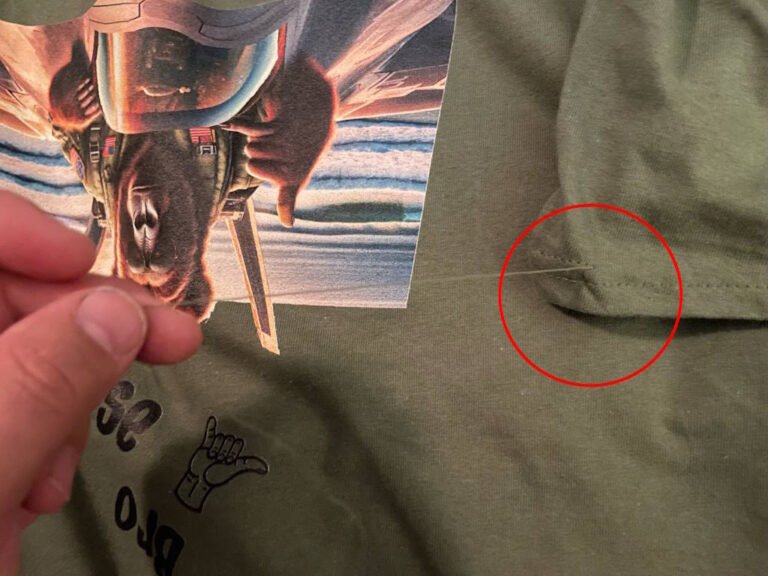 A close up image of a thread that came unsewn on the sleeve of a military green shirt, with a red circle highlighting the issue. There is a graphic image also in the picture. A hand is pulling the thread to make it visible in the picture.