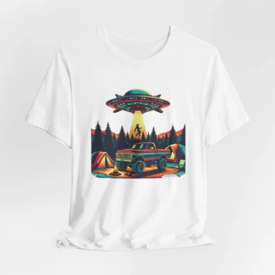 White colored t-shirt mockup of a camping scene, with a 1980 style pick-up truck, tents, and an alien space ship in the background, abducting a Big Foot looking creature from out of a pine forest. Colors are red, orange, blue, and yellow.