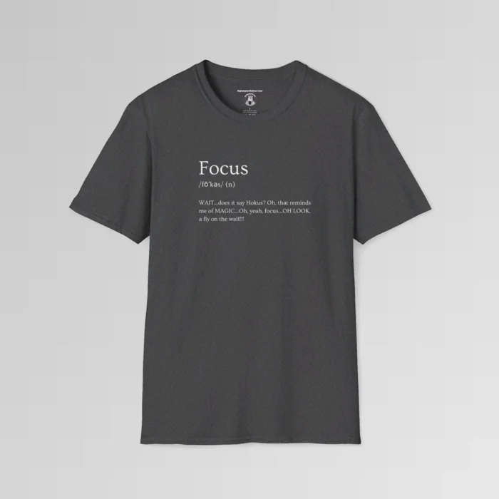 Dark heather t-shirt mockup with the word "Focus" on the front, in the appearance of a word in a dictionary, including pronunciation beneath the main word. The text beneath says "WAIT...does it say Hokus? Oh, that reminds me of MAGIC...Oh, yeah, focus...OH LOOK, a fly on the wall!!!!"