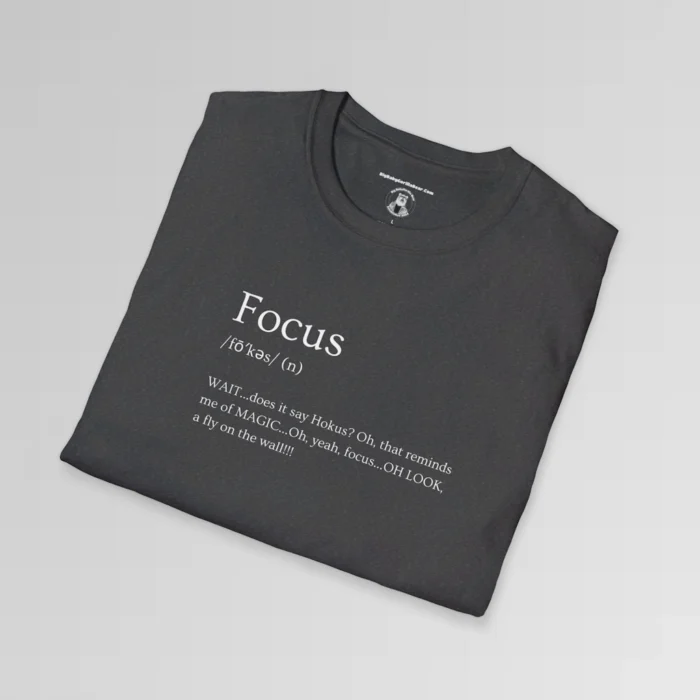 Dark heather folded t-shirt mockup with the word "Focus" on the front, in the appearance of a word in a dictionary, including pronunciation beneath the main word. The text beneath says "WAIT...does it say Hokus? Oh, that reminds me of MAGIC...Oh, yeah, focus...OH LOOK, a fly on the wall!!!!"