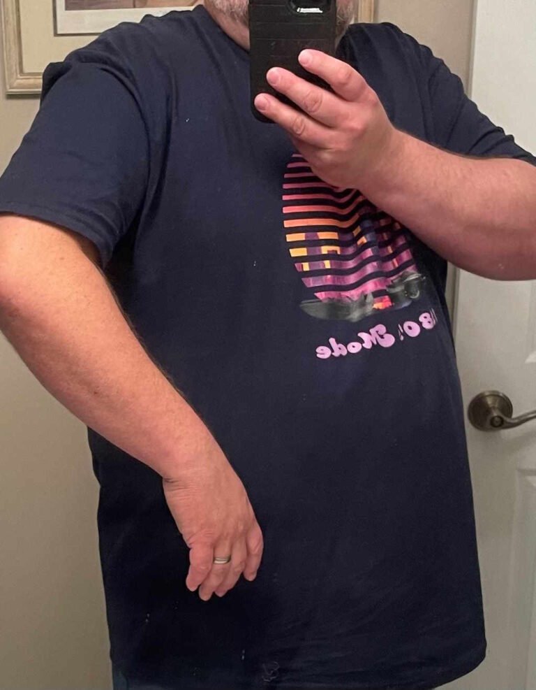 A picture of someone taking a picture in the mirror, raising one arm to show a defect in the fit of the sleeve of a t-shirt.