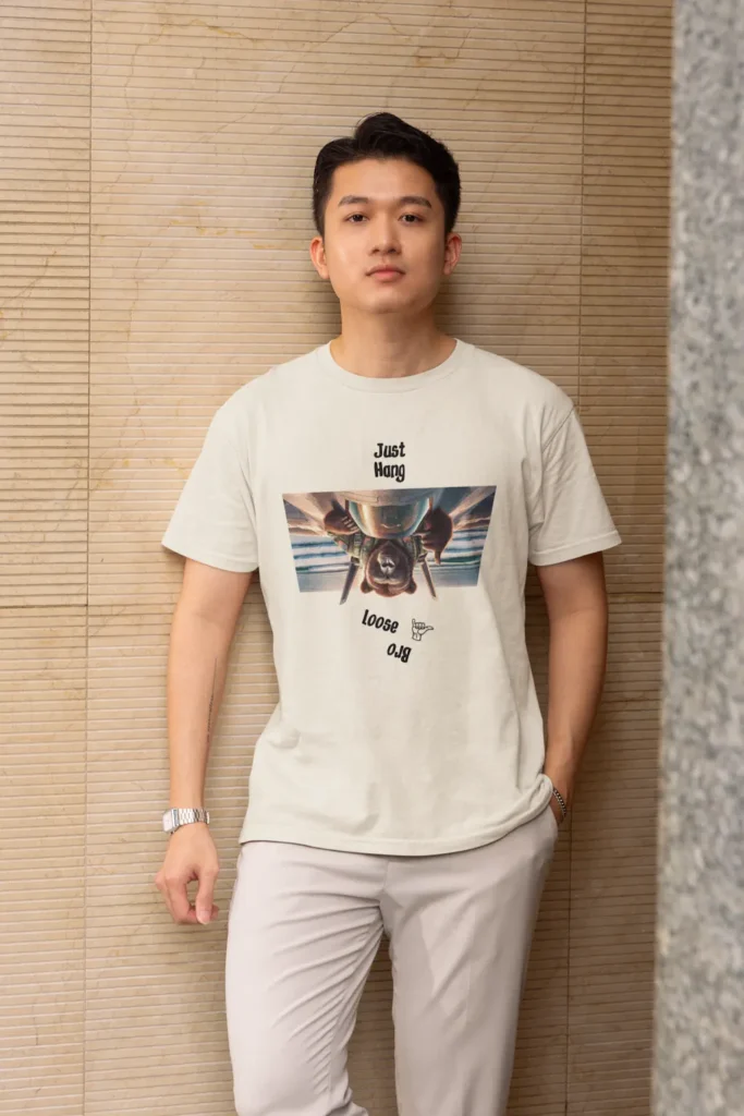 Young Asian male against a wall, wearing the "Just hang Loose Upside Down" Natural colored graphic tee.