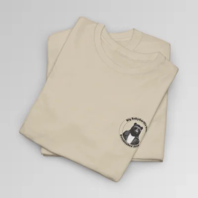 A sand colored folded t-shirt mockup with the Big Baby Gorilla Bear circular logo on the front left chest. The logo has the main mascot, a grizzly bear and gorilla hybrid, wearing jacket and shades, in the middle of the circle. On the outside, it has the company name on top and "Established 2024" on the bottom.