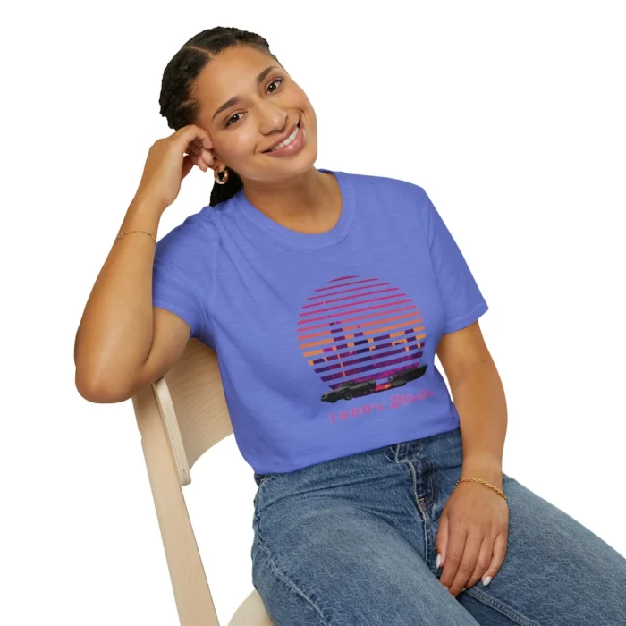 Attractive female sitting and wearing the Heather Royal colored "Miami 1980's Mode" tee, showcasing a city at sunset with a vertical blind effect, and a convertible car and speed boat at the bottom of the image, and "1980's Mode" written beneath the image in a retro font style.