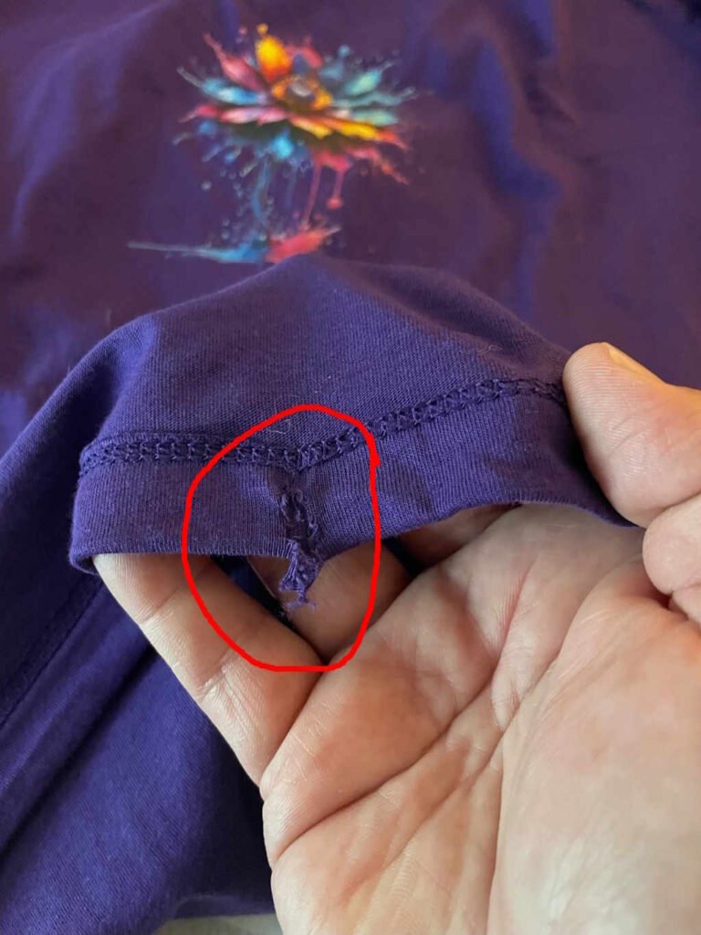 A hand holding the bottom of a purple t-shirt, with the main t-shirt graphic still visible. The image has a red circle highlighting a tear in the fabric.