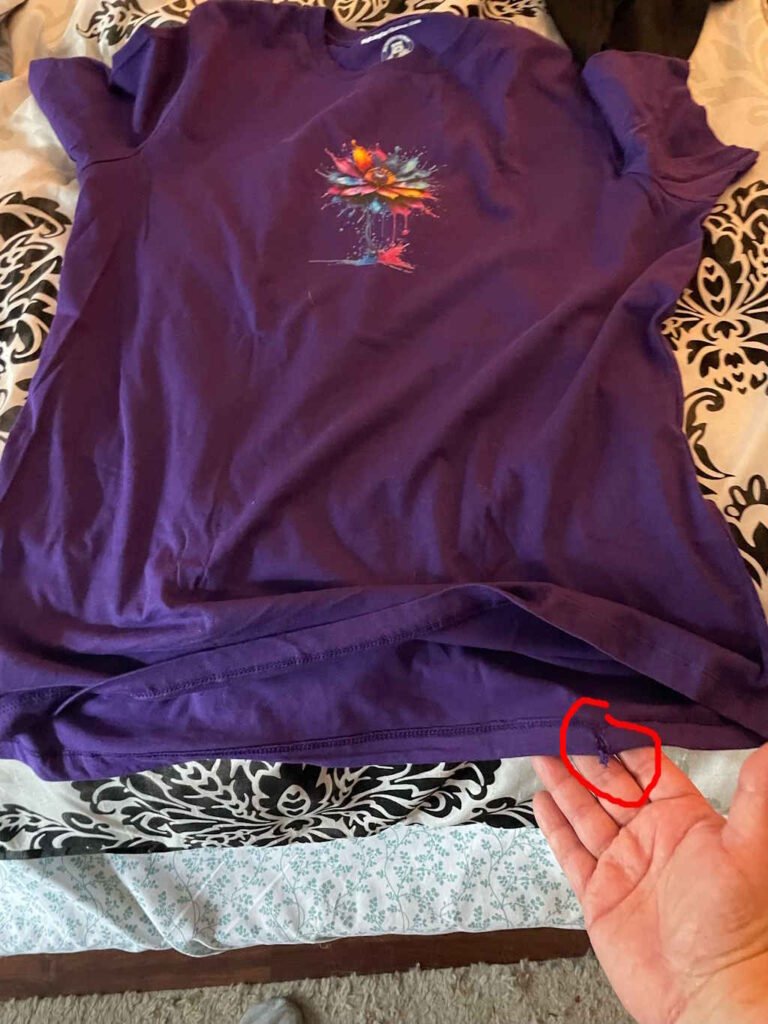 A far away shot of a purple t-shirt with a defect on the bottom of the shirt. The image is shot from a wider angle to show the entire shirt., with a red circle highlighting where the defect is on the overall shirt.