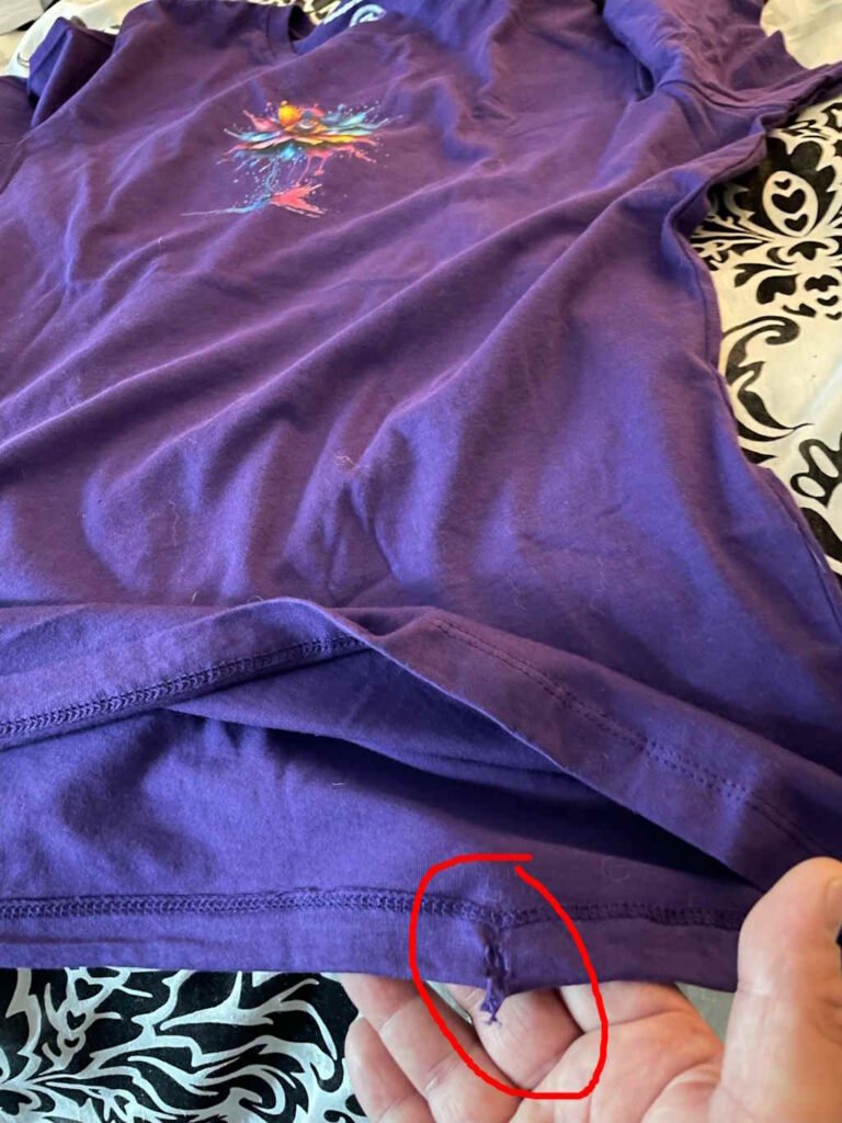 A hand holding the bottom of a purple t-shirt, with the main t-shirt graphic still visible. The image has a red circle highlighting a tear in the fabric. This shot is from a little wider angle.