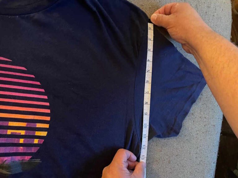 A close up of a tape measure measuring from the top of a sleeve stitch to the bottom of a sleeve stitch, where the shirt sleeve is sewn to the body of the t-shirt.