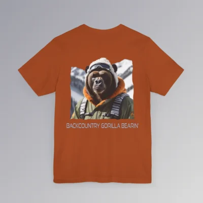 An autumn colored tee with a backside graphic of one of our iconic gorilla grizzly bears, fully dressed in an army/olive colored snow jacket with orange trim, white toboggan, and a backpack. There are snowy mountains in the background. Beneath the image is text that reads "Backcountry Gorilla Bearin'".