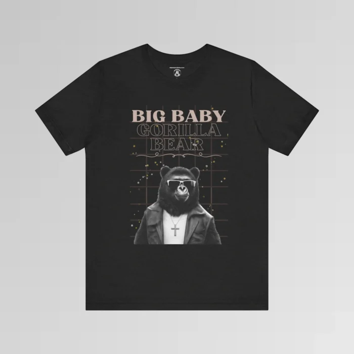 A black heather t-shirt mockup with our iconic grizzly bear and gorilla hybrid, wearing a leather jacket, cross necklace, and shades. The background of the shirt has a grid and some confetti. The words above the image are "Big Baby Gorilla Bear in two fonts, with a decorative ribbon separating the words from the mascot image. Retro look with tan, black, grey, and white colors.