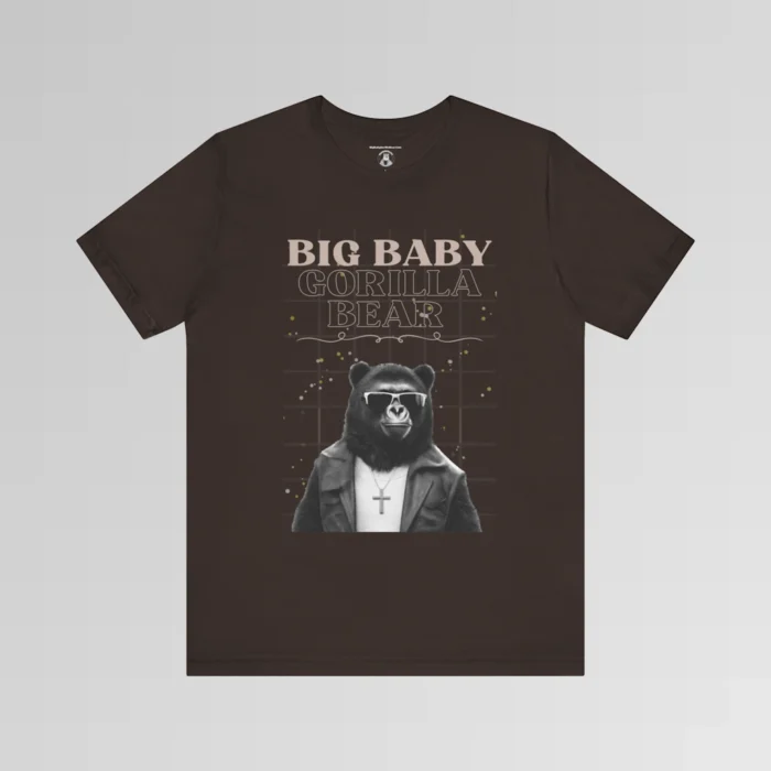 A brown t-shirt mockup with our iconic grizzly bear and gorilla hybrid, wearing a leather jacket, cross necklace, and shades. The background of the shirt has a grid and some confetti. The words above the image are "Big Baby Gorilla Bear in two fonts, with a decorative ribbon separating the words from the mascot image. Retro look with tan, black, grey, and white colors.