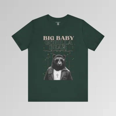 A forest green t-shirt mockup with our iconic grizzly bear and gorilla hybrid, wearing a leather jacket, cross necklace, and shades. The background of the shirt has a grid and some confetti. The words above the image are "Big Baby Gorilla Bear in two fonts, with a decorative ribbon separating the words from the mascot image. Retro look with tan, black, grey, and white colors.