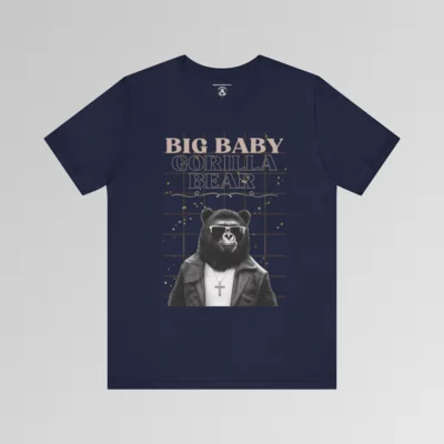 A navy t-shirt mockup with our iconic grizzly bear and gorilla hybrid, wearing a leather jacket, cross necklace, and shades. The background of the shirt has a grid and some confetti. The words above the image are "Big Baby Gorilla Bear in two fonts, with a decorative ribbon separating the words from the mascot image. Retro look with tan, black, grey, and white colors.