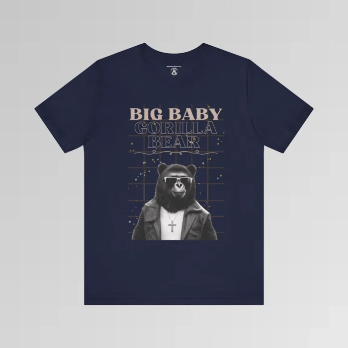 A navy t-shirt mockup with our iconic grizzly bear and gorilla hybrid, wearing a leather jacket, cross necklace, and shades. The background of the shirt has a grid and some confetti. The words above the image are "Big Baby Gorilla Bear in two fonts, with a decorative ribbon separating the words from the mascot image. Retro look with tan, black, grey, and white colors.