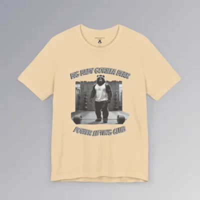 A soft cream colored tee with an image of one of our iconic gorilla-bear hybrid creatures in a tank top, in a gym, with the image in grayscale color scheme. 3D print is above and below the image. On top, it says "Big Baby Gorilla Bear", and on bottom it says "Power Lifting Club". Graphic on front.