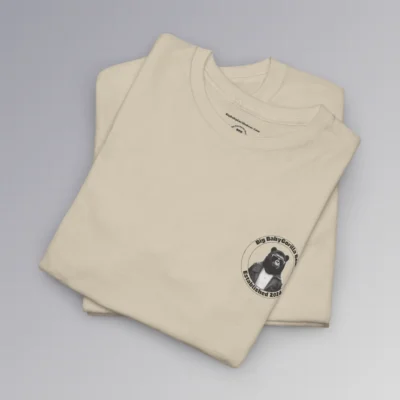 A folded, sand colored t-shirt with the Big Baby Gorilla Bear Logo on the front. The logo is circular, with a circle inside a circle, Inside the circles is "Big Baby Gorilla Bear" on the top and :"Established 2024" on the bottom. In the center of the circle, is our mascot, a grizzly-gorilla bear character, with a leather jacket, and shades, and a gold cross chain.