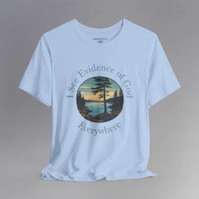 A baby blue t-shirt with a cove on a lake, and a slightly off centered focal point pine tree, with mountains in the distance, and pine trees on the edge of the circular graphic. The sun is setting in the image. Text around the image says "I See Evidence of God" on the top half, and "Everywhere" on the bottom half; text wrapped around the image.