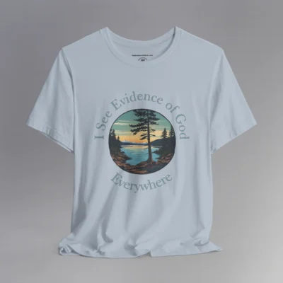 A light blue t-shirt with a cove on a lake, and a slightly off centered focal point pine tree, with mountains in the distance, and pine trees on the edge of the circular graphic. The sun is setting in the image. Text around the image says "I See Evidence of God" on the top half, and "Everywhere" on the bottom half; text wrapped around the image.