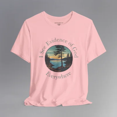 A pink t-shirt with a cove on a lake, and a slightly off centered focal point pine tree, with mountains in the distance, and pine trees on the edge of the circular graphic. The sun is setting in the image. Text around the image says "I See Evidence of God" on the top half, and "Everywhere" on the bottom half; text wrapped around the image.