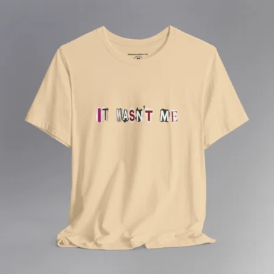 A soft cream tee with magazine cutout letters of various fonts and colors arranged to spell out "It Wasn't Me", on the front of the t-shirt.
