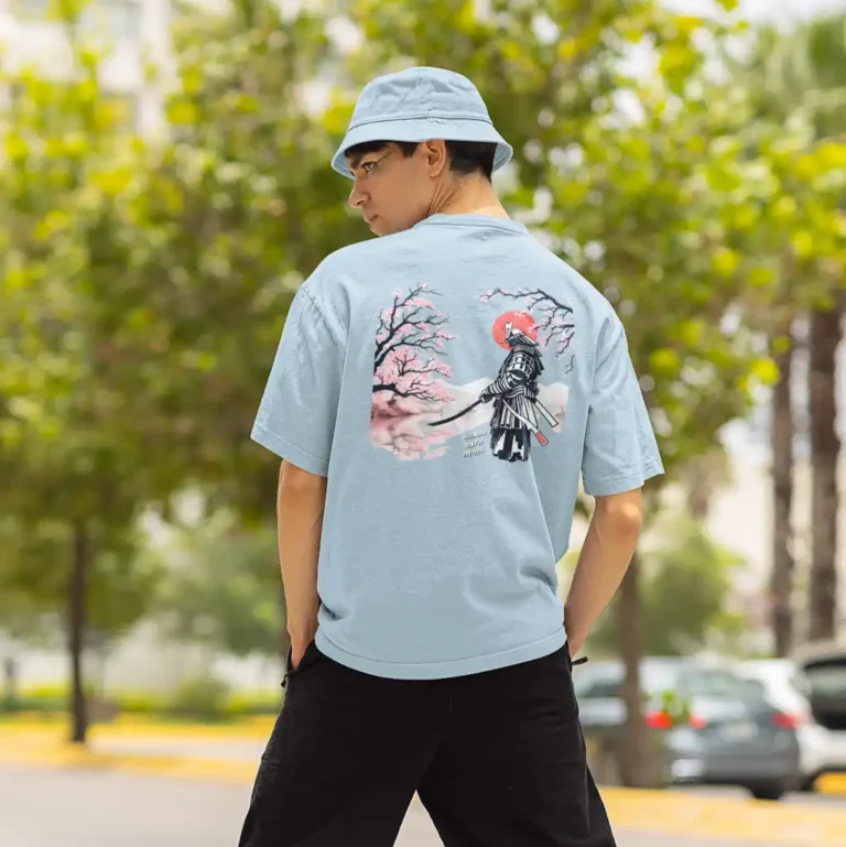 A man wearing a streetwear outfit in a public space. Blue hat and shirt, with black pants. The shirt has a cherry blossom/samurai image on the back.