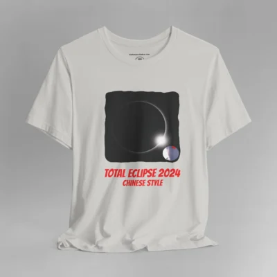 A silver colored graphic tee with the 2024 Total Eclipse image, but with the twist of the Chinese Spy Balloon flying in the lower right corner of the iamge. In red, there is writing beneath that says "Total Eclipse 2024" in a bigger font, and then "Chinese Style" in a smaller font beneath the other writing.