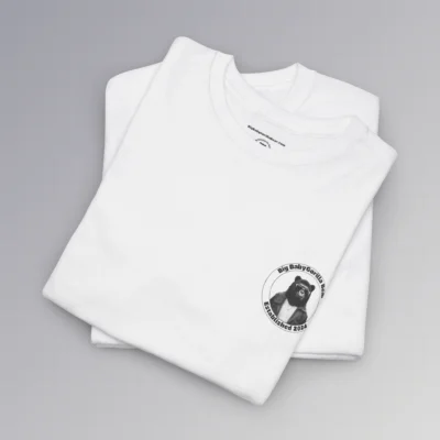 Frontside of a folded white colored tee, with the circular Big Baby gorilla Bear 2024 logo, with two circles, and on the inside, the image of our iconic mascot in leather jacket and shades. Between the inner circles of the logo, on the top is the brand name, and on the bottom it says "established 2024". The "Kiss The Sun Breakdancing" T-shirt.