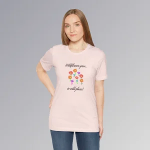 A female model wearing a soft pink colored t-shirt with blue, red, orange, and purple coneflower-like flowers, with green and mauve colored stems. In a handwriting font, is says "Wildflowers grow..." above the flowers, and "in wild places!" beneath the flowers.