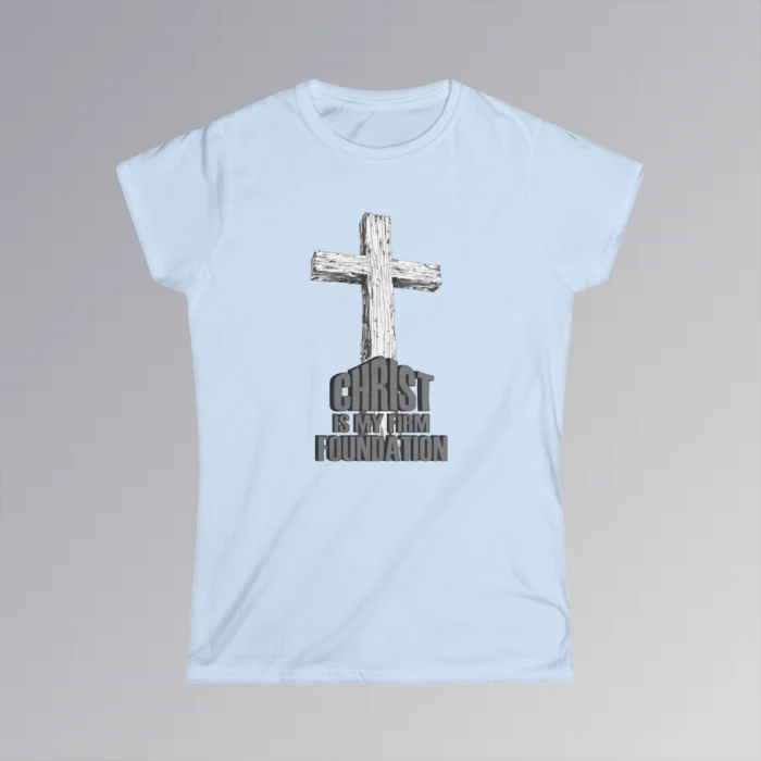 The front of a t-shirt with 3D text which states "Christ Is My Firm Foundation", from a slightly left perspective, in the shape of a simple house, with an old, wooden cross in the background. Color: Light Blue. Her Style.