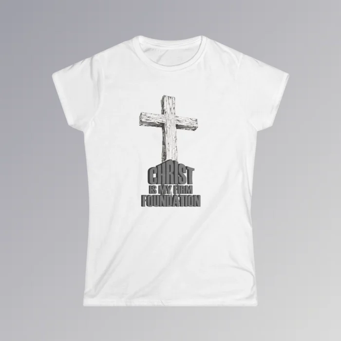 The front of a t-shirt with 3D text which states "Christ Is My Firm Foundation", from a slightly left perspective, in the shape of a simple house, with an old, wooden cross in the background. Color: White. Her Style.