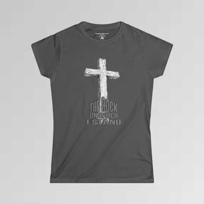 The front of a t-shirt with 3D text which states "The Rock On Which I Stand", from a slightly left perspective, in the shape of a simple house, with an old, wooden cross in the background. Color: Charcoal. Her Style.