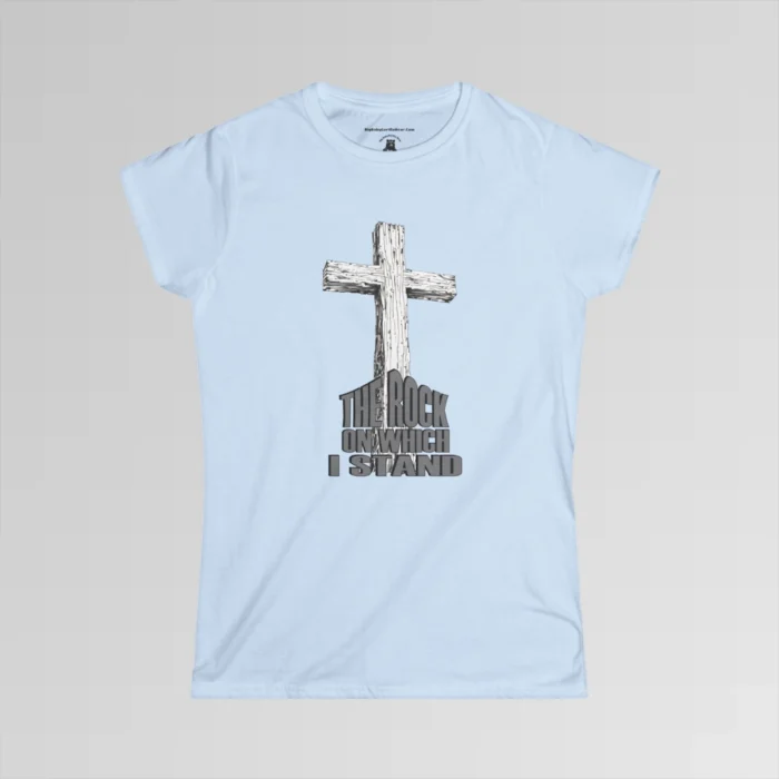 The front of a t-shirt with 3D text which states "The Rock On Which I Stand", from a slightly left perspective, in the shape of a simple house, with an old, wooden cross in the background. Color: Light Blue. Her Style.