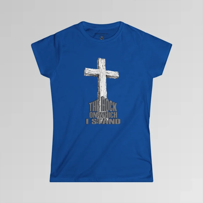 The front of a t-shirt with 3D text which states "The Rock On Which I Stand", from a slightly left perspective, in the shape of a simple house, with an old, wooden cross in the background. Color: Royal. Her Style.