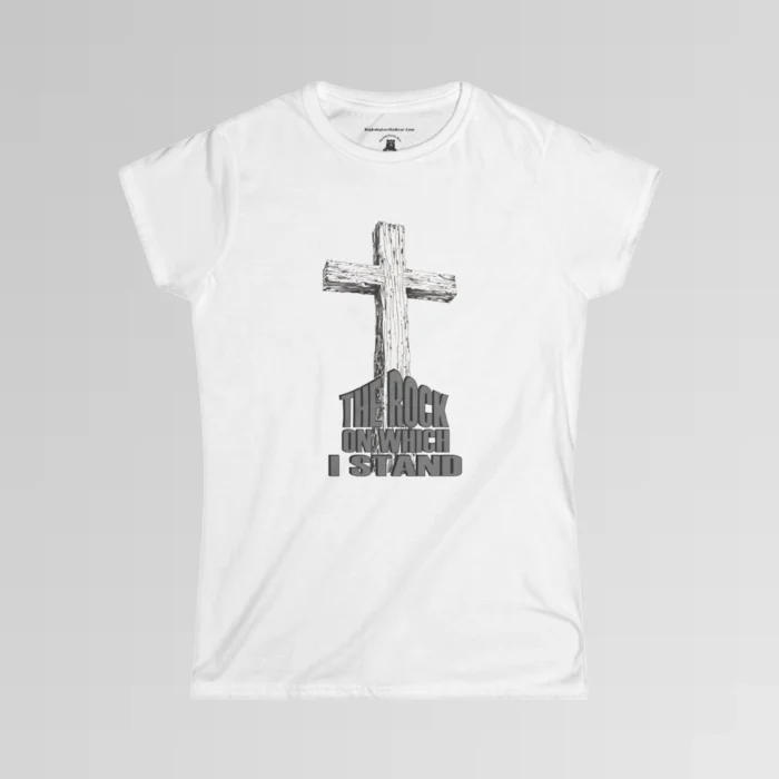 The front of a t-shirt with 3D text which states "The Rock On Which I Stand", from a slightly left perspective, in the shape of a simple house, with an old, wooden cross in the background. Color: White. Her Style.