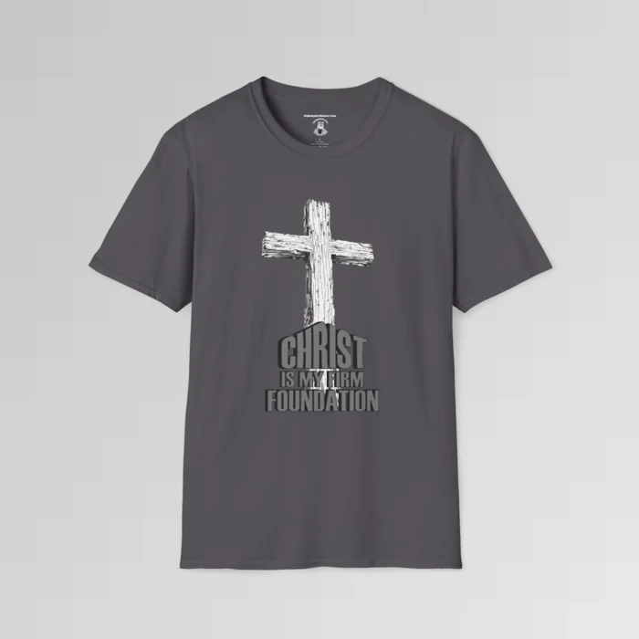 The front of a t-shirt with 3D text which states "Christ Is My Firm Foundation", from a slightly left perspective, in the shape of a simple house, with an old, wooden cross in the background. Color: Charcoal. His Style.