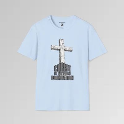 The front of a t-shirt with 3D text which states "Christ Is My Firm Foundation", from a slightly left perspective, in the shape of a simple house, with an old, wooden cross in the background. Color: Light Blue. His Style.