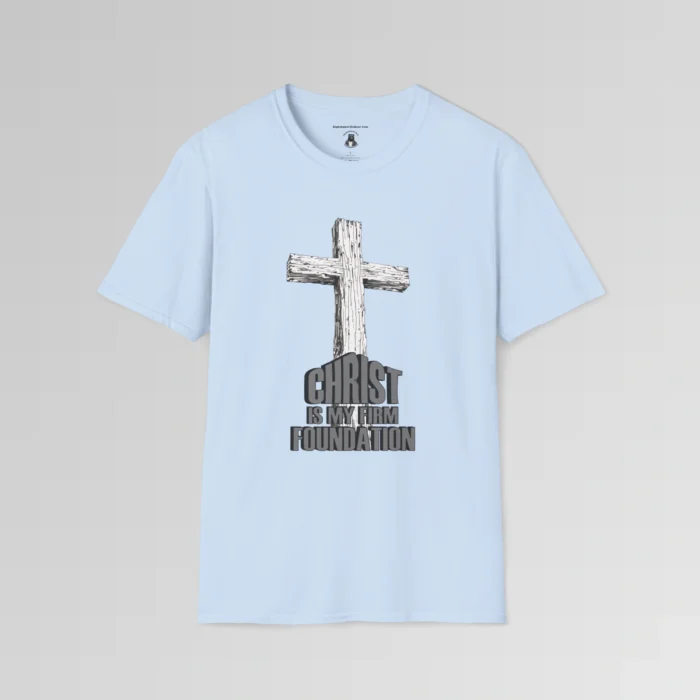 The front of a t-shirt with 3D text which states "Christ Is My Firm Foundation", from a slightly left perspective, in the shape of a simple house, with an old, wooden cross in the background. Color: Light Blue. His Style.