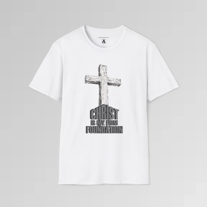 The front of a t-shirt with 3D text which states "Christ Is My Firm Foundation", from a slightly left perspective, in the shape of a simple house, with an old, wooden cross in the background. Color: White. His Style.