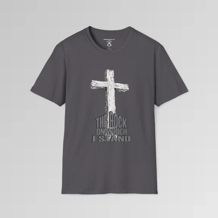 The front of a t-shirt with 3D text which states "The Rock On Which I Stand", from a slightly left perspective, in the shape of a simple house, with an old, wooden cross in the background. Color: Charcoal. His Style.