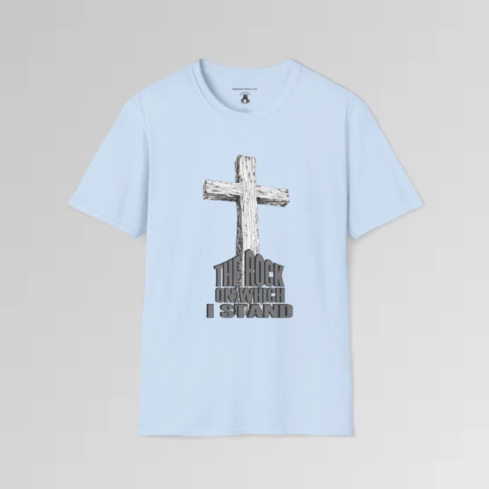 The front of a t-shirt with 3D text which states "The Rock On Which I Stand", from a slightly left perspective, in the shape of a simple house, with an old, wooden cross in the background. Color: Light Blue. His Style.