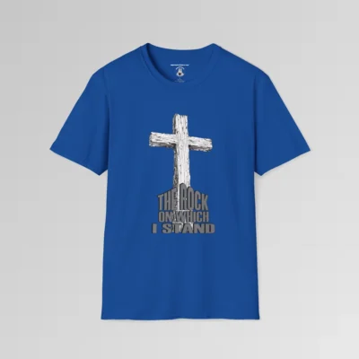 The front of a t-shirt with 3D text which states "The Rock On Which I Stand", from a slightly left perspective, in the shape of a simple house, with an old, wooden cross in the background. Color: Royal. His Style.