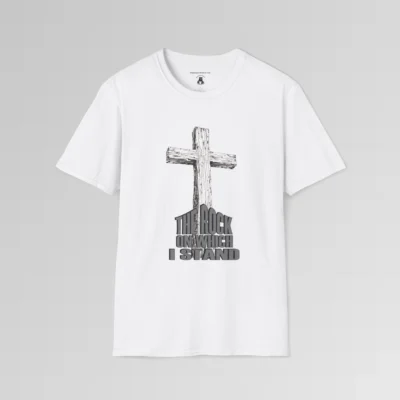 The front of a t-shirt with 3D text which states "The Rock On Which I Stand", from a slightly left perspective, in the shape of a simple house, with an old, wooden cross in the background. Color: White. His Style.
