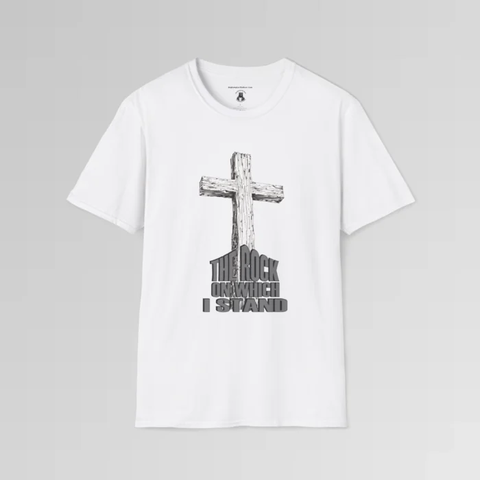 The front of a t-shirt with 3D text which states "The Rock On Which I Stand", from a slightly left perspective, in the shape of a simple house, with an old, wooden cross in the background. Color: White. His Style.