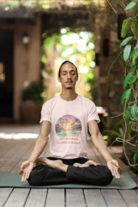 Male model in a sitting yoga pose, with a serene background, wearing a soft pink tee, with a sunset beach scene, and the 3D words "My Vacay Goes Hard", with the font slightly curved.