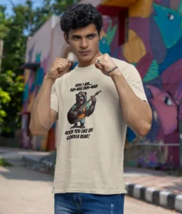 A guy in a fighting pose, wearing the sand colored Rock You Like a gorilla Bear T-shirt by Big Baby Gorilla Bear. The shirt has a gorilla bear on it in shorts, tee, and button up, wearing red shades, and playing a red electric guitar. Words on the shirt say "Here I am duh-nah duh-nah rock you like a gorilla bear!