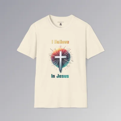 A natural colored t-shirt with the words "I Believe" and "In Jesus" above and below a Christian cross, with a gradient white to very light blue filling the cross.. Around the cross is a vibrant circle with lines like stars shooting out, in the colors ranging from yellow to orange, pink, to teal and blue. The words "I Believe" has a gradient fill color of yellow to orange. The words "In Jesus" has a gradient fill color of teal to dark teal/blue.