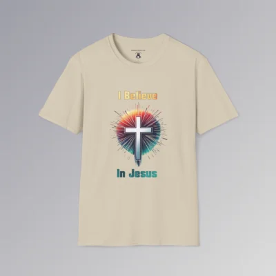 A sand colored t-shirt with the words "I Believe" and "In Jesus" above and below a Christian cross, with a gradient white to very light blue filling the cross.. Around the cross is a vibrant circle with lines like stars shooting out, in the colors ranging from yellow to orange, pink, to teal and blue. The words "I Believe" has a gradient fill color of yellow to orange. The words "In Jesus" has a gradient fill color of teal to dark teal/blue.