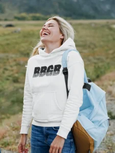 A female model wearing the BBGB Simple Hoodie, White, while walking in nature, laughing, and carrying a bookbag (light blue).