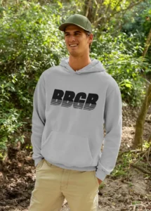 A male model wearing the BBGB Simple Hoodie, Sport Grey, while standing in nature, smiling, and wearing a green hat and khaki pants.