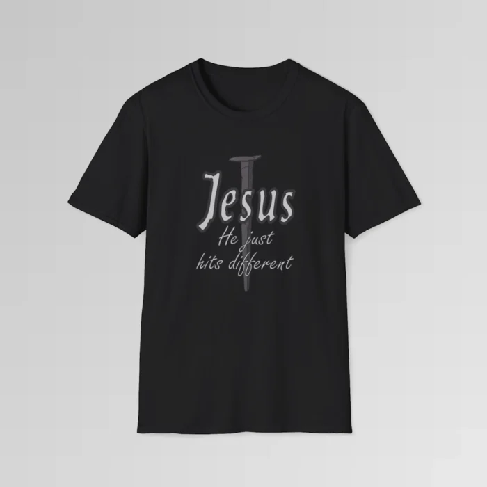Black colored softstyle tee, with a crucifixion spike, and the name "Jesus" in big print over top of it. Beneath Jesus, it says "He just hits different", in a cursive font. The font is silver, with a dark grey border.